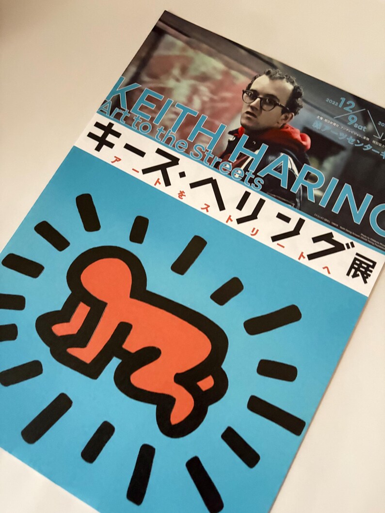 May include: A blue poster with a black and red illustration of a baby in a circle of black rays. The poster advertises a Keith Haring exhibition titled "Art to the Streets" with the text "Keith Haring Art to the Streets" in white and "キース・ヘリング展" in red.