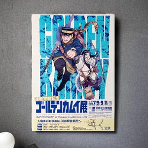 May include: A poster for the "Golden Kamuy Special Exhibition" with two illustrated characters holding weapons. The poster is blue and white, with Japanese text and the dates "7/9-9/11 2022".