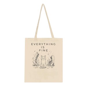 May include: A beige canvas tote bag featuring a black line art illustration of a cat sitting calmly in the center of a ruined city landscape. Above the cat, the text reads EVERYTHING IS FINE in a simple, clean, black sans-serif typeface.