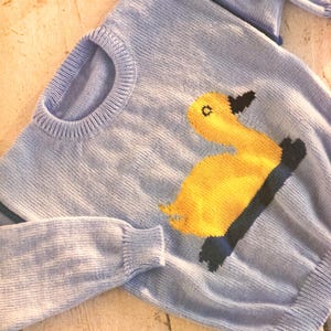 May include: A light blue knit sweater featuring a large yellow duck design with black accents. The sweater has a crew neck and long sleeves. The duck has a black beak and eye detail. The sweater is made of a soft, knitted material.