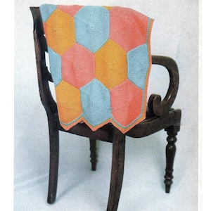以下が含まれることがあります： 青、オレンジ、ピンクの色合いの幾何学模様が施されたニットのベビーブランケット。ブランケットは木製の椅子の背もたれにかけられています。