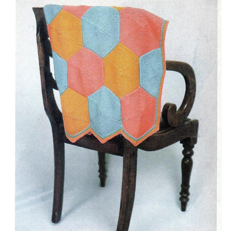 Op de afbeelding: Een gebreid babydekentje met een geometrisch patroon in tinten blauw, oranje en roze. Het dekentje is over de rugleuning van een houten stoel gelegd.