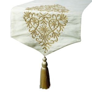 May include: A white table runner with a gold embroidered design and a gold tassel.