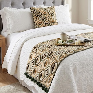 May include: A bed with white bedding, a gray button-tufted headboard, and a decorative geometric patterned throw. A matching pillow and a tray with a mug, candles, and a magazine are on the bed. The throw has green pom-poms.