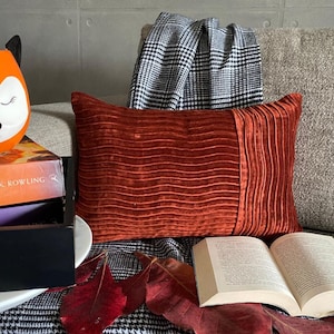 May include: A red velvet cushion with a pleated design sits on a black and white checked throw blanket. The cushion is on a sofa with a book and autumn leaves on the blanket.