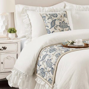 May include: A white bedding set featuring a ruffled duvet cover and matching pillow shams. A decorative throw pillow and a long bed runner are included, both displaying a blue and beige floral paisley pattern on a cream-colored background.
