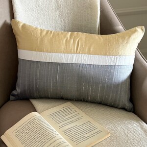 May include: A rectangular throw pillow featuring a horizontal colour-blocked design with a golden yellow top section, a narrow white middle stripe, and a dark grey bottom section. The fabric has a visible slubbed texture throughout the surface.