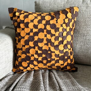 Pode incluir: Almofada decorativa quadrada com um padr&atilde;o de c&iacute;rculos castanhos e laranja alternados. A almofada est&aacute; colocada num sof&aacute; cinzento com uma manta a condizer. O design adiciona um toque de textura e cor ao ambiente de tons neutros.