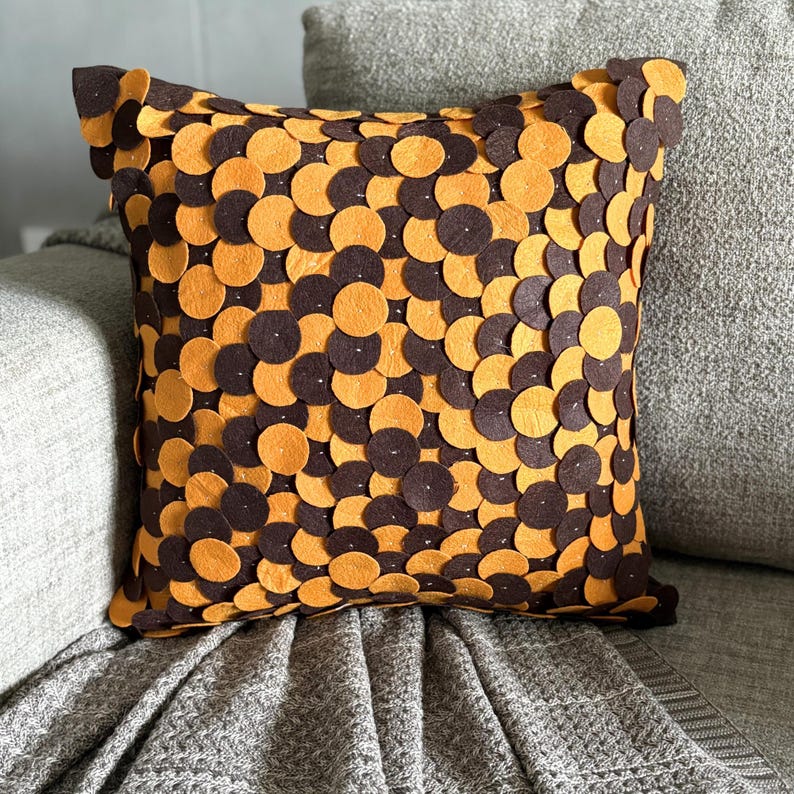 Pode incluir: Almofada decorativa quadrada com um padr&atilde;o de c&iacute;rculos castanhos e laranja alternados. A almofada est&aacute; colocada num sof&aacute; cinzento com uma manta a condizer. O design adiciona um toque de textura e cor ao ambiente de tons neutros.