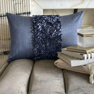 May include: A rectangular throw cushion in a slate blue fabric featuring a wide, central vertical band of dark blue gathered fabric ruffles. The cushion has a textured, woven appearance and is positioned behind a stack of books and a small box.