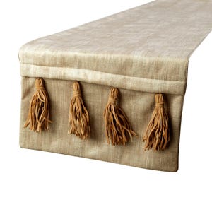 May include: A rectangular table runner in a light tan, textured fabric. The end of the runner features a horizontal stitched seam and four evenly spaced, dangling tassels in a matching tan color, each composed of multiple thin fabric strips.