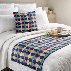 Puede incluir: Un cojín cuadrado y un camino de cama rectangular con un patrón geométrico de formas entrelazadas en tonos azul marino, rojo, amarillo y crema. Los artículos textiles se muestran sobre una cama blanca con ropa de cama blanca.