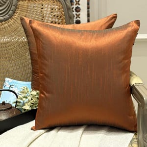 May include: Two square decorative throw pillows with a rich, copper-colored fabric. The pillows have a subtle vertical stripe pattern and are placed on a chair with a woven back. The pillows add a touch of warmth and elegance to the space.