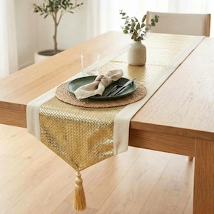 Pode incluir: Um caminho de mesa metálico dourado com um padrão de grelha texturizado e bordas de tecido cor de creme. O caminho apresenta uma borla decorativa na extremidade e está colocado sobre uma mesa de madeira com um lugar americano redondo.