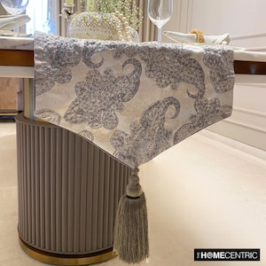 May include: A white table runner with a grey paisley pattern and a gold tassel. The runner is on a table with a grey base and gold trim.