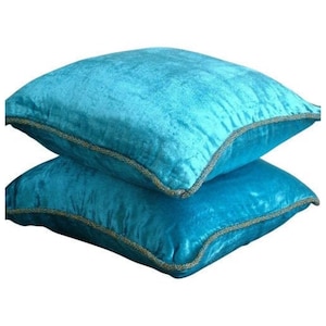May include: Two turquoise blue velvet throw pillows with gold trim. The pillows are stacked on top of each other.