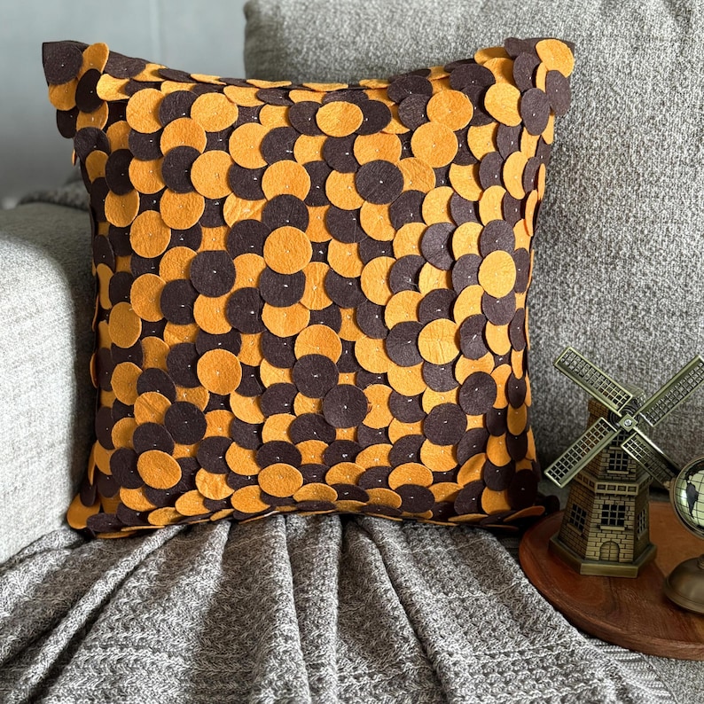 Pode incluir: Almofada quadrada decorativa com um padr&atilde;o de c&iacute;rculos castanhos e laranja. A almofada est&aacute; colocada num sof&aacute; cinzento e numa manta cinzenta. A almofada tem uma superf&iacute;cie texturizada.