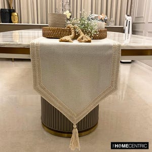 May include: A white and gray herringbone table runner with a beige crocheted trim and tassels. The runner is on a marble table with a brown pedestal base.