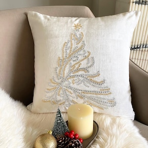 May include: A white decorative pillow with an embroidered Christmas tree design in gold and silver beads. The pillow is on a beige chair, with a candle, ornaments, and pinecones in front.