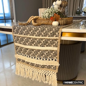 May include: A brown and beige table runner with a geometric pattern and fringe trim. The runner is on a white table with a gold trim. The table runner is decorated with a wooden bead garland and a wicker basket filled with white flowers and pumpkins.