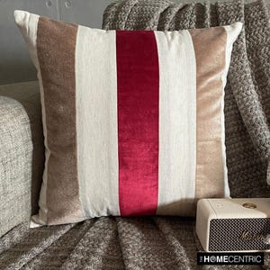 Puede incluir: Una almohada decorativa con un patrón a rayas en beige, blanco y burdeos. La almohada está hecha de terciopelo y tiene un tacto suave y lujoso.