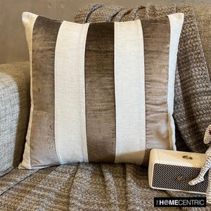May include: A brown and white striped throw pillow with a velvet texture. The pillow is sitting on a brown couch with a blanket.