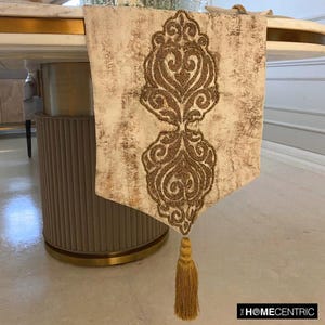 Puede incluir: Un camino de mesa blanco con un diseño de cuentas doradas y una borla dorada. El camino de mesa tiene un aspecto desgastado y es perfecto para agregar un toque de elegancia a cualquier mesa.