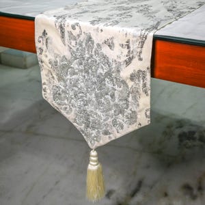 May include: A cream-coloured table runner featuring a prominent silver glitter damask pattern. The fabric has a pointed end finished with a single, matching cream-coloured tassel. The runner is draped over the edge of a wooden table surface.