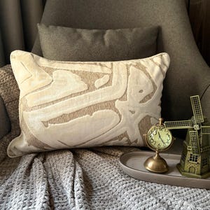 May include: A rectangular throw cushion featuring a cream-coloured velvet-like abstract pattern over a textured beige fabric base. The cushion is positioned next to a small brass-toned analogue clock and a miniature metal windmill figurine on a tray.