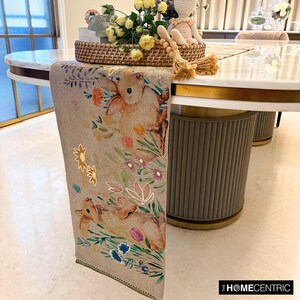Op de afbeelding: Een beige tafelkleed met een bloemenpatroon met konijnen en bloemen. De loper ligt op een witte tafel met een gouden rand.