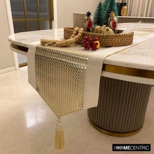 May include: A rectangular table runner featuring a central panel of metallic gold woven material flanked by off-white fabric borders. The runner includes a decorative beige tassel at the pointed end and is styled with a rattan tray containing nutcrackers.