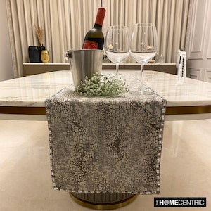 Peut inclure: Un chemin de table rectangulaire avec un motif texturé en peau de serpent argenté et taupe, bordé de perles. Il est posé sur une table en marbre blanc avec un seau à glace en argent, une bouteille de vin et deux verres à vin.