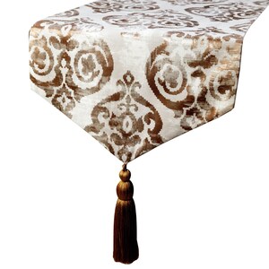 May include: A rectangular table runner featuring a white base with a repeating brown damask pattern. The fabric has a slight sheen and terminates in a pointed end finished with a long, multi-tiered brown tassel hanging from the center point.