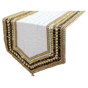 Op de afbeelding: Een witte tafelloper met een geplooid middenpaneel en decoratieve randen. De zijkanten zijn versierd met rijen goudkleurige kralen, metallic zilveren lovertjes, gele kristallen en donkerbruine vierkante tegeltjes in een geometrisch patroon.