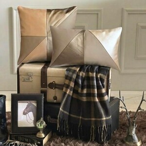 May include: Two decorative throw pillows featuring a geometric colour-blocked design in shades of tan, beige, and metallic gold. One pillow is square and the other is rectangular, both displaying a smooth, satin-like finish with clean seams.