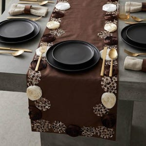 May include: A brown table runner decorated with white and brown fabric rosettes and circular sequin clusters. The runner is set with matte black plates, gold-toned forks, knives, and spoons, alongside beige linen napkins with wooden napkin rings.