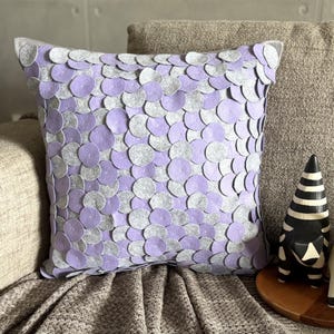 May include: A square throw pillow featuring a dense, layered pattern of overlapping circular felt discs in alternating shades of light purple and soft grey. The circular pieces are attached to the front surface, creating a textured, scale-like appearance across the entire face of the cushion.