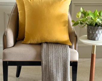May include: Two square mustard yellow velvet throw pillows are placed on a beige upholstered chair. A grey knit throw blanket with a textured pattern is draped over the seat of the chair, which features black legs and a sleek, modern frame design.