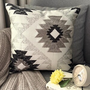 May include: Square cushion with a geometric design in black, grey, and white. The cushion has a central diamond shape and triangle accents, with beaded embroidery. The cushion is on a grey sofa.