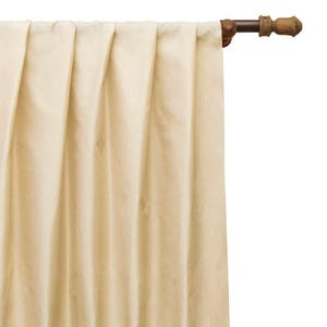 May include: A solid cream-colored curtain panel with vertical pleats hanging from a dark brown wooden curtain rod. The fabric has a smooth, matte texture and hangs in uniform folds, extending downward from the rod with a simple, clean finish.
