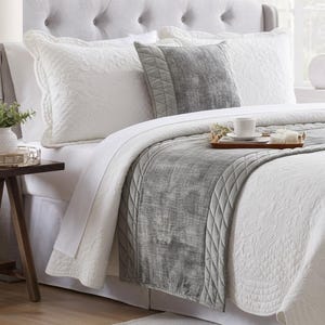 Op de afbeelding: Een wit doorgestikt sprei-set bestaande uit een textuurrijke witte sprei met ingewikkelde stiksels, bijpassende kussenslopen, een contrasterende grijze fluwelen bedloper met ruitvormig doorgestikte randen en een bijpassend grijs sierkussen.