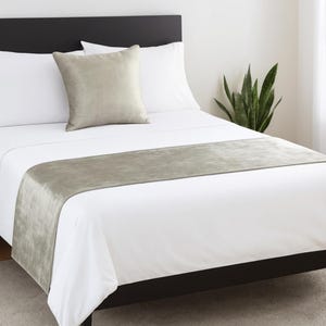 Puede incluir: Un juego de cama que incluye una funda nórdica blanca, dos almohadas blancas y un cojín cuadrado de terciopelo verde salvia. Un camino de cama rectangular a juego, también de terciopelo verde salvia, se extiende horizontalmente a los pies.