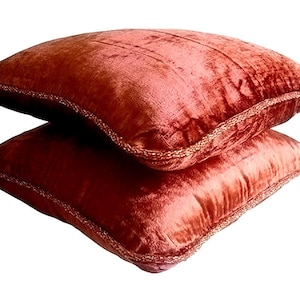 May include: Two red velvet cushion covers with a decorative trim. The cushions are stacked on top of each other.