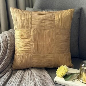May include: A square throw pillow in a solid golden-tan color featuring a textured, crinkled fabric surface. The pillow is constructed with a grid-like pattern of stitched panels, creating a distinct block design across the front face.