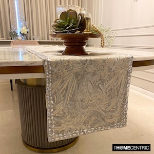 May include: A rectangular table runner in a light grey and silver tone featuring an intricate, abstract textured pattern. The edges are embellished with a border of clear, faceted crystals. The brand name THE HOMECENTRIC is printed in the bottom corner.