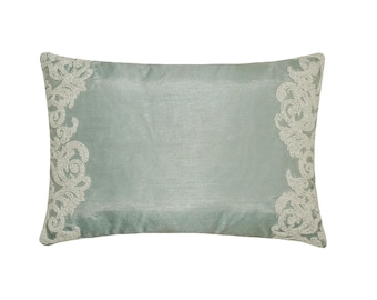 May include: A rectangular throw pillow in a soft sage green color with a smooth, slightly lustrous finish. The pillow features intricate, cream-colored embroidered scrollwork patterns along the left and right vertical edges of the front.