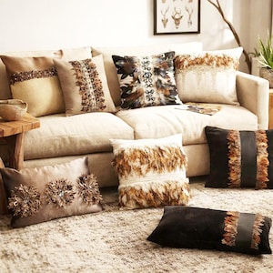 May include: A collection of decorative throw pillows in various shapes and sizes, featuring textured fabrics, brown and black color palettes, and embellishments including layered feathers, sequins, and central bands on a beige sofa.