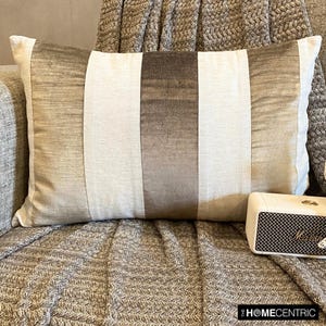 May include: A beige and brown striped throw pillow with a subtle sheen. The pillow is sitting on a brown and gray textured throw blanket.