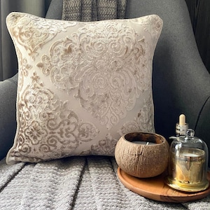 Puede incluir: Una almohada beige con un patrón de damasco se sienta en un sofá gris con una manta de lana gris. Un portavelas de cáscara de coco y una campana de vidrio con una vela dentro están en una bandeja de madera frente a la almohada.