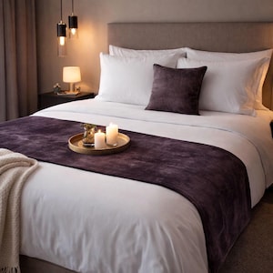 May include: A bed featuring white linens, a dark purple velvet bed runner, and a matching dark purple square throw pillow. A round gold tray sits on the bed, holding two lit white pillar candles and a small glass vase with greenery.